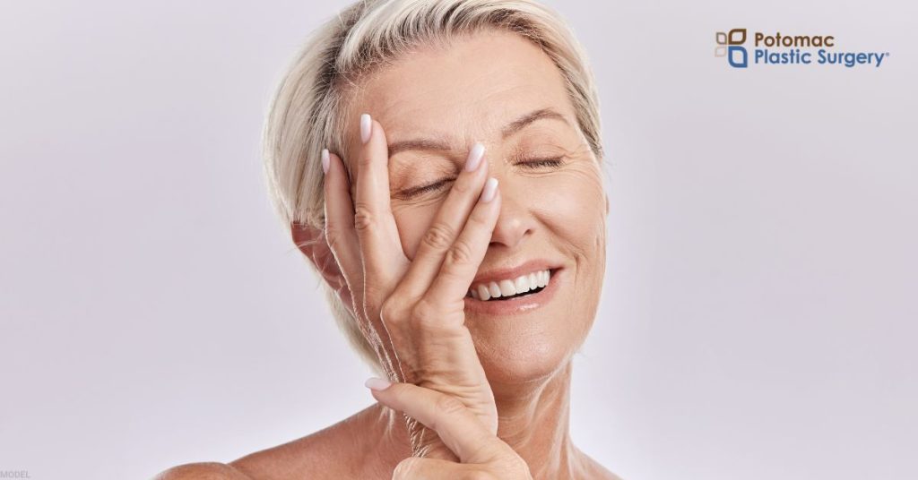 Mature woman smiling and gently touches her face. (MODEL) Potomac Plastic Surgery Logo